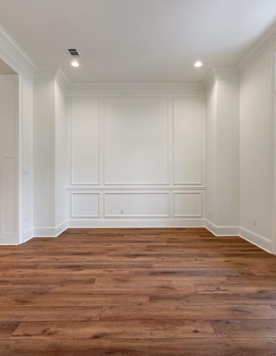 Modern, empty room with hardwood floors, white walls with wainscoting, and a view into a kitchen with white cabinets.