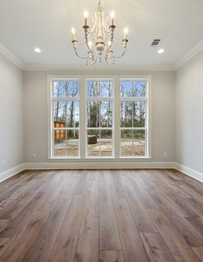 An empty room with hardwood flooring, large windows with a view of leafless trees, and a chandelier.