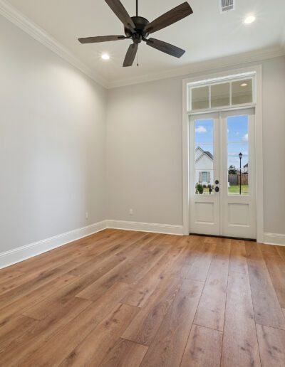 Empty room with wooden flooring, white walls, and a ceiling fan, leading to an open door with a view outside.