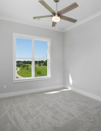 An empty room with gray carpet, white walls, and a ceiling fan, featuring a window with a view of the outdoors.