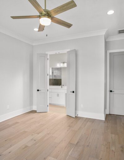 A clean, empty room with hardwood floors, white walls, and a ceiling fan, featuring a view into an adjoining room with a bathroom vanity.