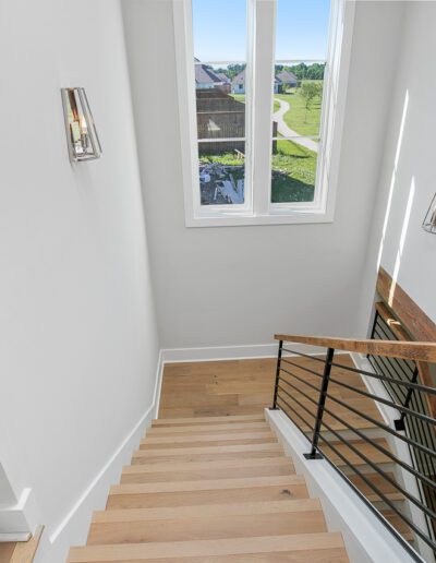 Modern staircase with wooden steps, metal balusters, and wall-mounted lights, overlooking a residential neighborhood through a tall window.