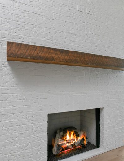 A modern interior featuring a white painted brick fireplace with a wooden mantel and a fire burning, flanked by tall windows on a sunny day.