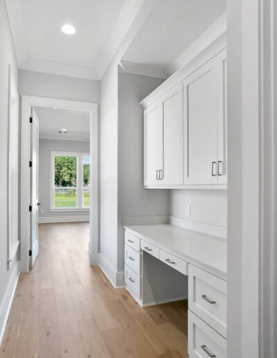 Modern hallway in a home with built-in white cabinets and workspace, leading to a well-lit room with a view of greenery outside.