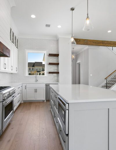 Bright modern kitchen with white cabinets, stainless steel appliances, and an island, leading to an adjacent living space with a staircase.